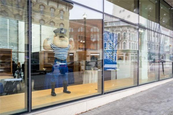 Laker Mascot Louie the Laker stands in a window flexing his biceps for passersby. 