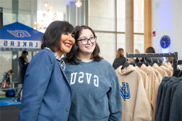 A women in a GVSU sweatshirt, and a woman in a suit jacket pose together looking toward a camera that is to the left out of frame. 