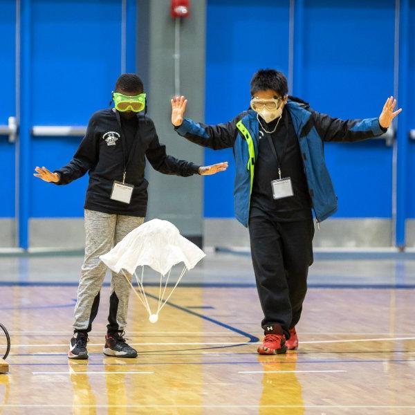 Two people wearing goggles hold their hands in the air as they watch a small parachute lift off from a gymnasium floor.