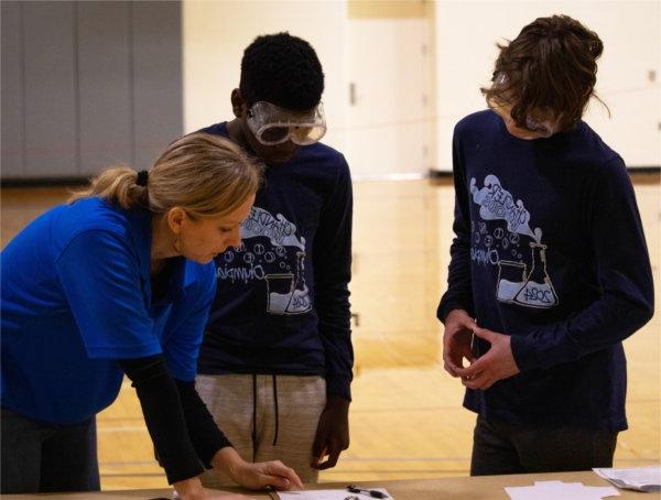 two students look at a table with goggles on, a coach is at right in a blue shirt