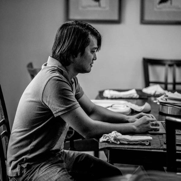 a black and white photo of John Wen typing on a laptop at his dining room table; he is wearing a golf shirt