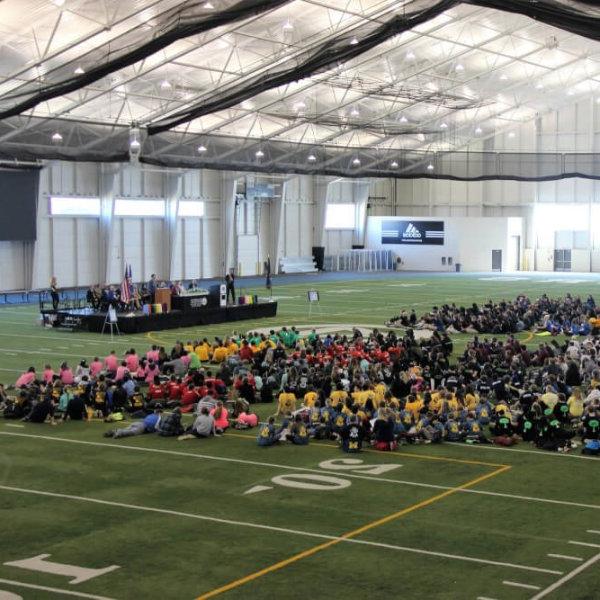 A large group of people sits on a floor with football-like yard markers. There also is a podium where people are sitting and one person is talking.